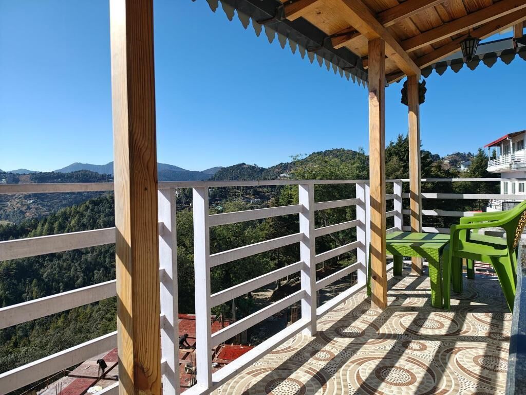 Un balcón con vistas a un valle y montañas, barandilla de madera y sillas de plástico verde.