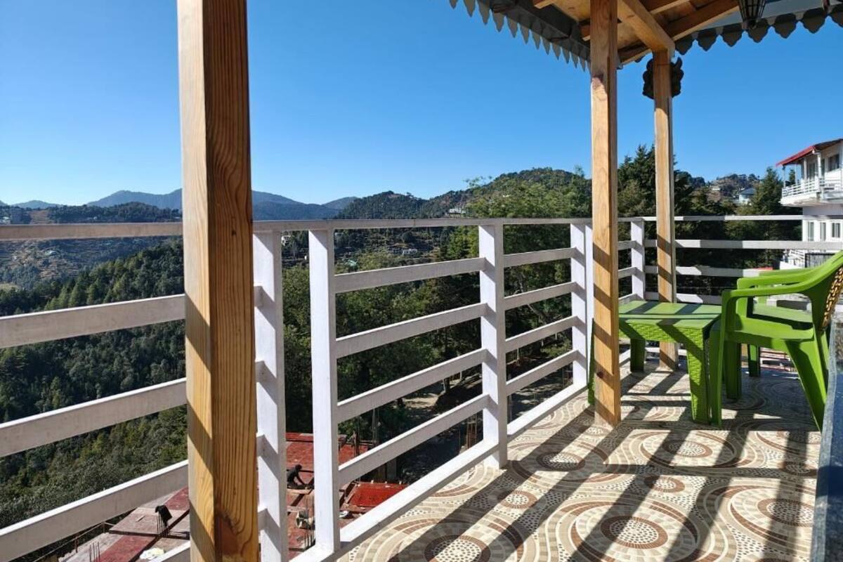 Un balcon donnant sur une vallée et des montagnes, avec une balustrade en bois et des chaises en plastique vertes.