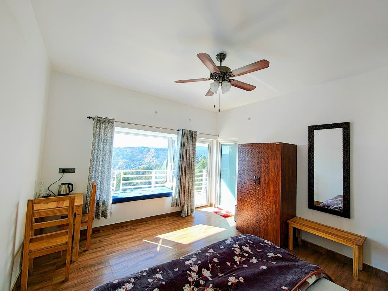 Un dormitorio con cama, ventilador de techo, armario de madera, un banco, ventana con cortinas y vistas al exterior.