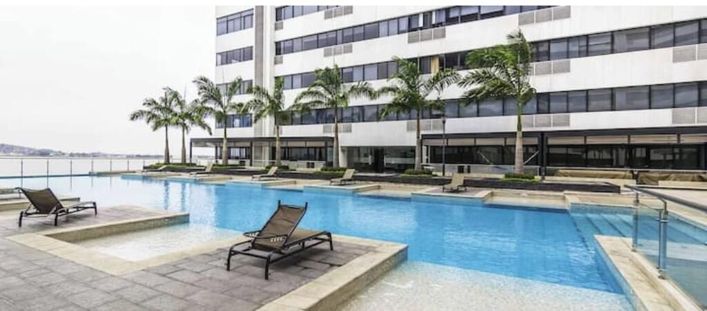A swimming pool with lounge chairs, palm trees, and a modern building with multiple floors.