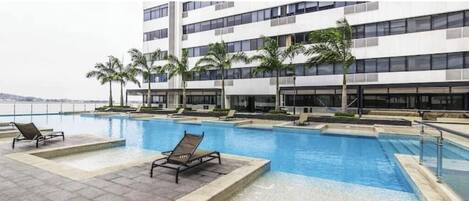 A swimming pool with lounge chairs, palm trees, and a modern building with multiple floors.