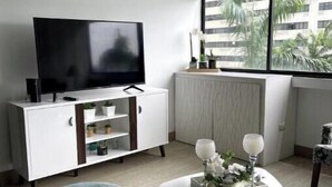 Ein modernes Wohnzimmer mit einem Flachbildfernseher, einer weißen Kommode, einem runden Couchtisch mit Dekoration und einem Fenster mit Blick auf Palmen.