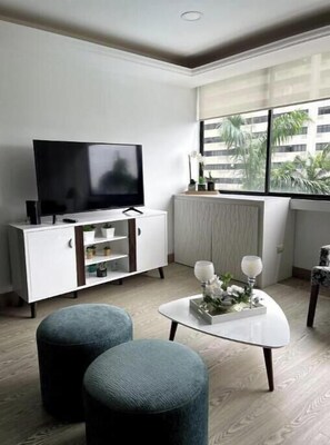 Ein modernes Wohnzimmer mit einem Flachbildfernseher, einer weißen Kommode, einem runden Couchtisch mit Dekoration und einem Fenster mit Blick auf Palmen.