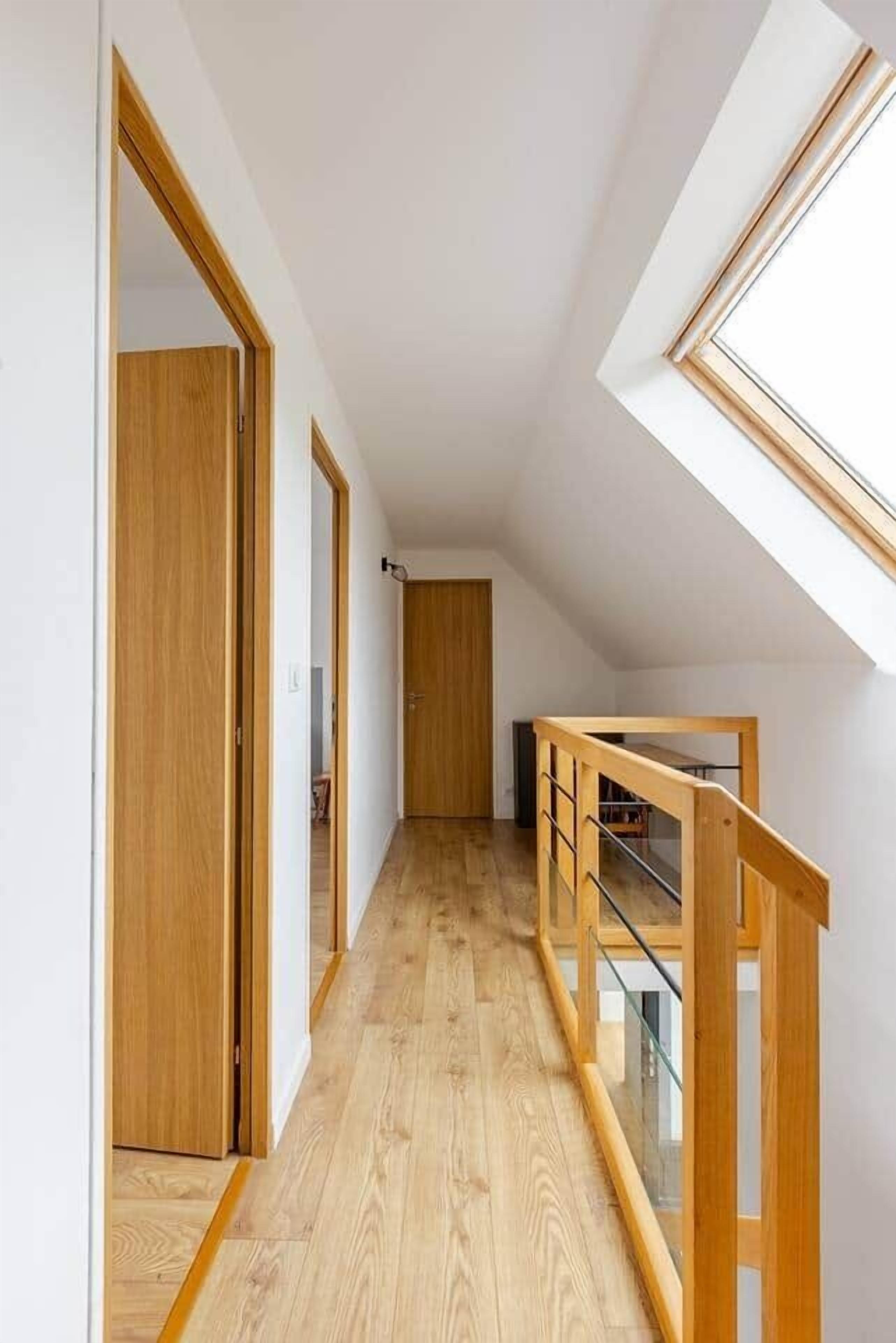 A narrow hallway with wooden flooring and doors, leading to a room with a glass railing and a sloped ceiling.