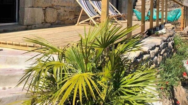 Een gebouw van steen met een houten terras, twee lounge fauteuils en een potplant.