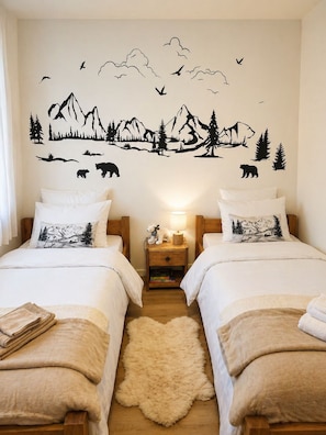 Ein Schlafzimmer mit zwei Betten, einem Nachttisch mit Lampe, einem flauschigen Teppich und einer Wanddekoration mit Berglandschaft.
