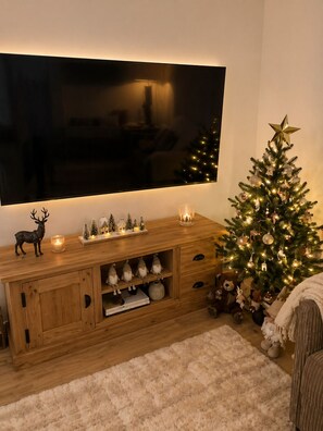 Ein gemütliches Wohnzimmer mit einem großen Flachbildfernseher an einem Holzschrank, einem Weihnachtsbaum mit Lichterkette und verschiedenen Dekoartikeln.