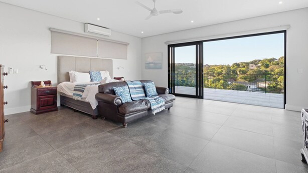 A spacious bedroom with a large bed, a sofa, a nightstand, and a sliding glass door leading to a balcony with a city view. - Room-tuna Room - Staynest Guesthouse (Ballito)