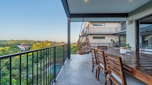 A balcony with a dining table and chairs, a hanging chair, and a view of a building and greenery. - Room-tuna Room - Staynest Guesthouse (Ballito)