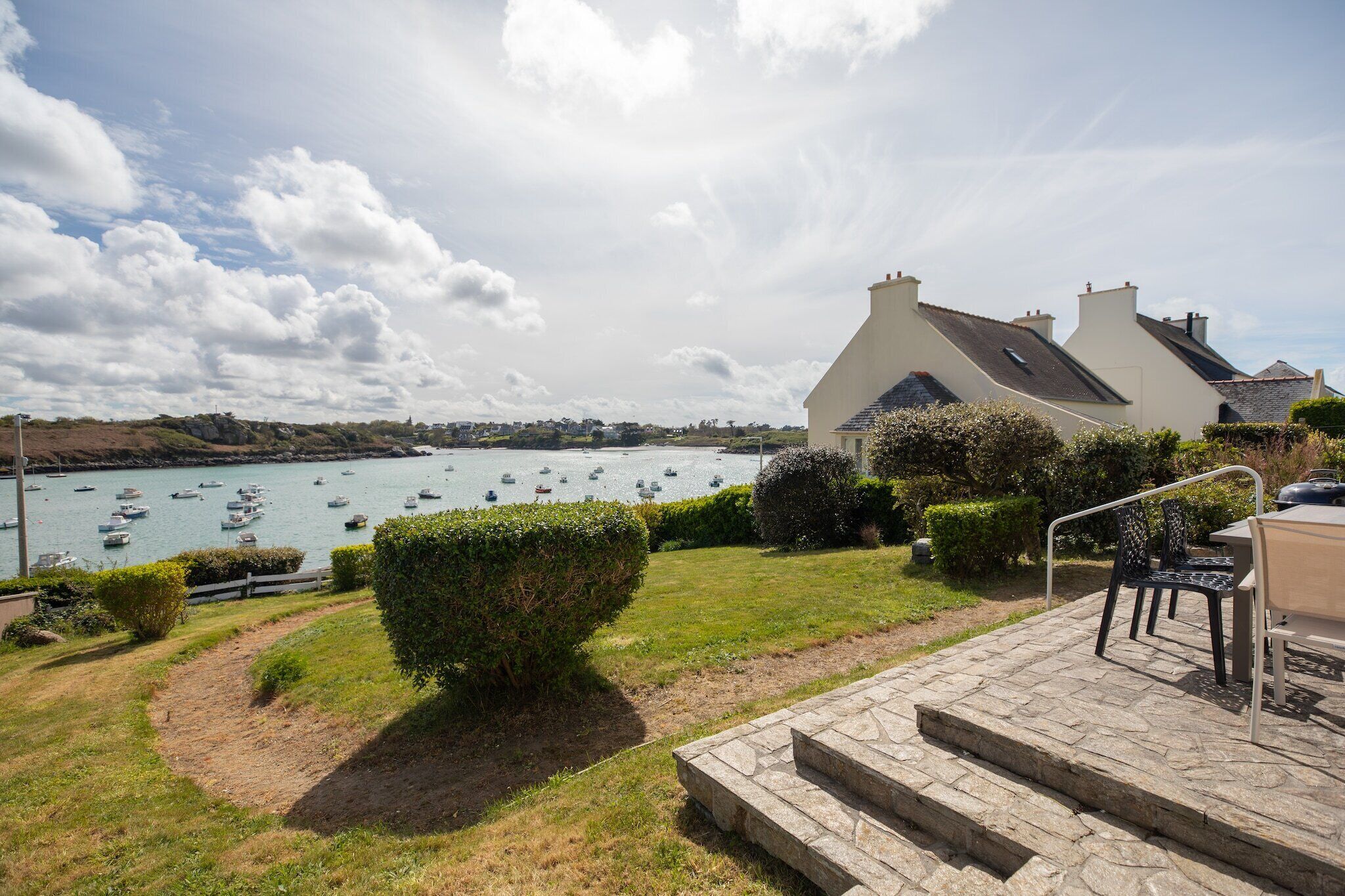 A coastal property with a view of boats on the water, a paved patio with outdoor seating, and a stone staircase leading to the entrance.