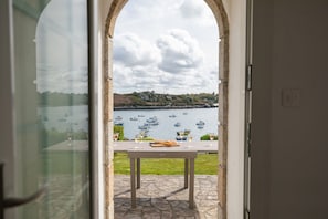Vista a través de un arco hacia una mesa con vasos y una baguette, con vista a un cuerpo de agua con barcos.