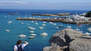 Paesaggio costiero con imbarcazioni, uno scoglio in primo piano e un edificio con tetto piatto.