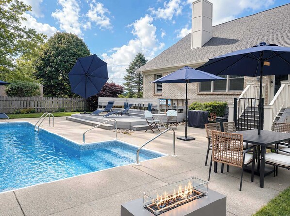 A swimming pool with lounge chairs and umbrellas, a fire pit, and a brick building in the background.