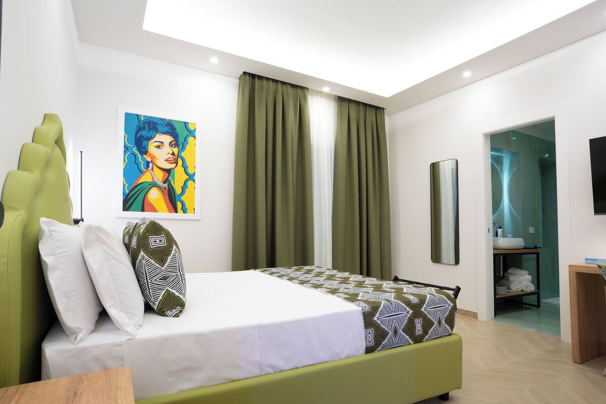 Ett modernt hotellrum med en säng, gröna gardiner, en tavla och ett badrum som syns genom en öppen dörr.