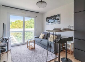 A modern living room with a sofa, a dining table with chairs, a large window, and a flat-screen TV. - Appartement t2 4 couchages et parking (La Rochelle)