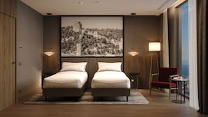 A hotel room with a double bed, two bedside lamps, a red chair, a small table, and a large cityscape painting on the wall.