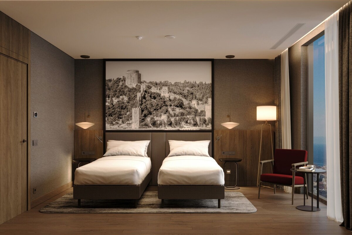 A hotel room with a double bed, two bedside lamps, a red chair, a small table, and a large cityscape painting on the wall.