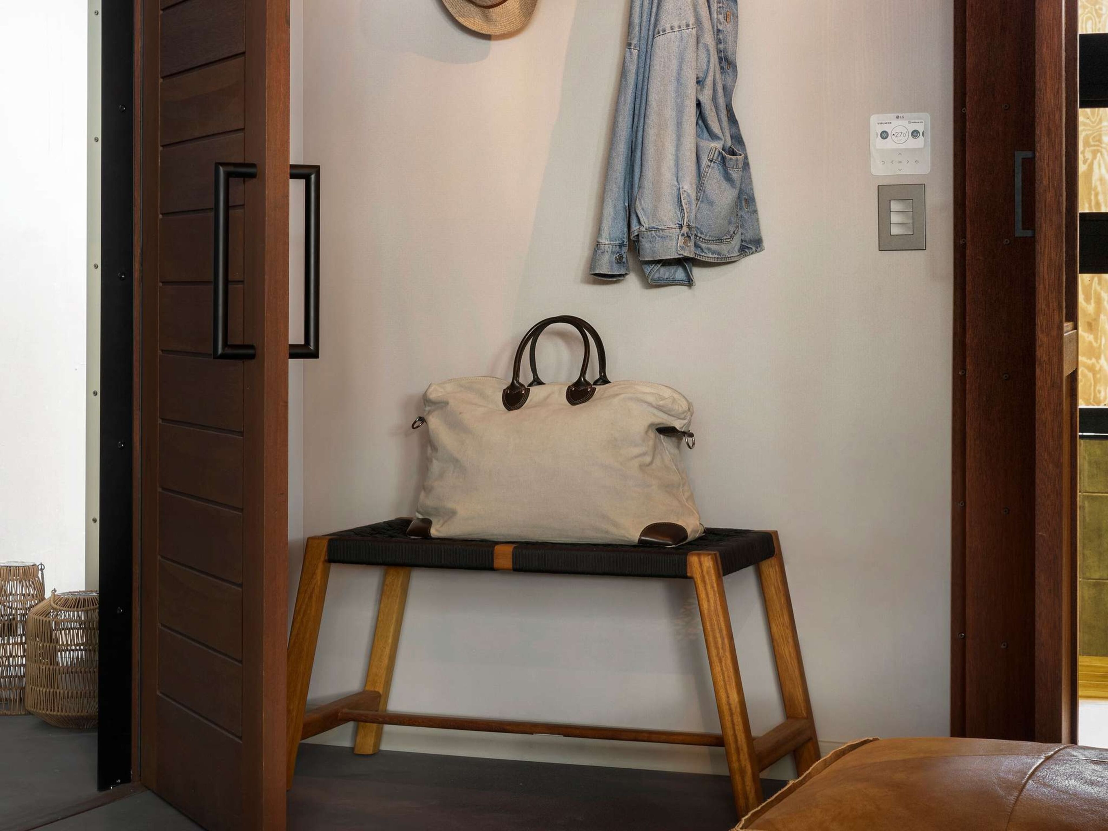 Une pièce avec une porte en bois, un chapeau accroché au mur, une veste en jean, un sac à main beige posé sur un tabouret en bois et un thermostat.