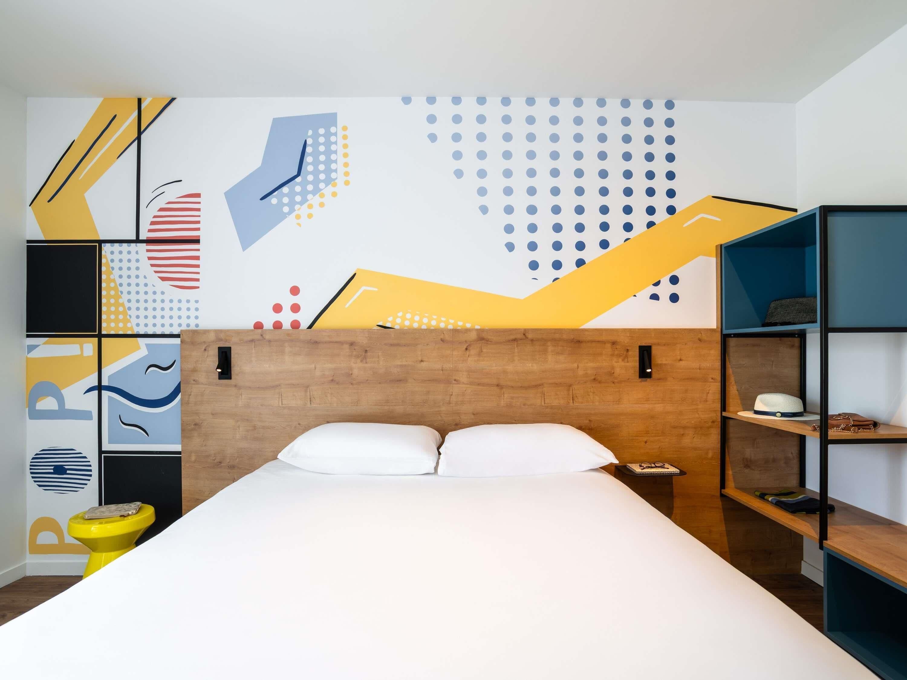Un dormitorio moderno con cabecera de madera, cama con sábanas blancas, inodoro amarillo y una pared decorada con arte abstracto.