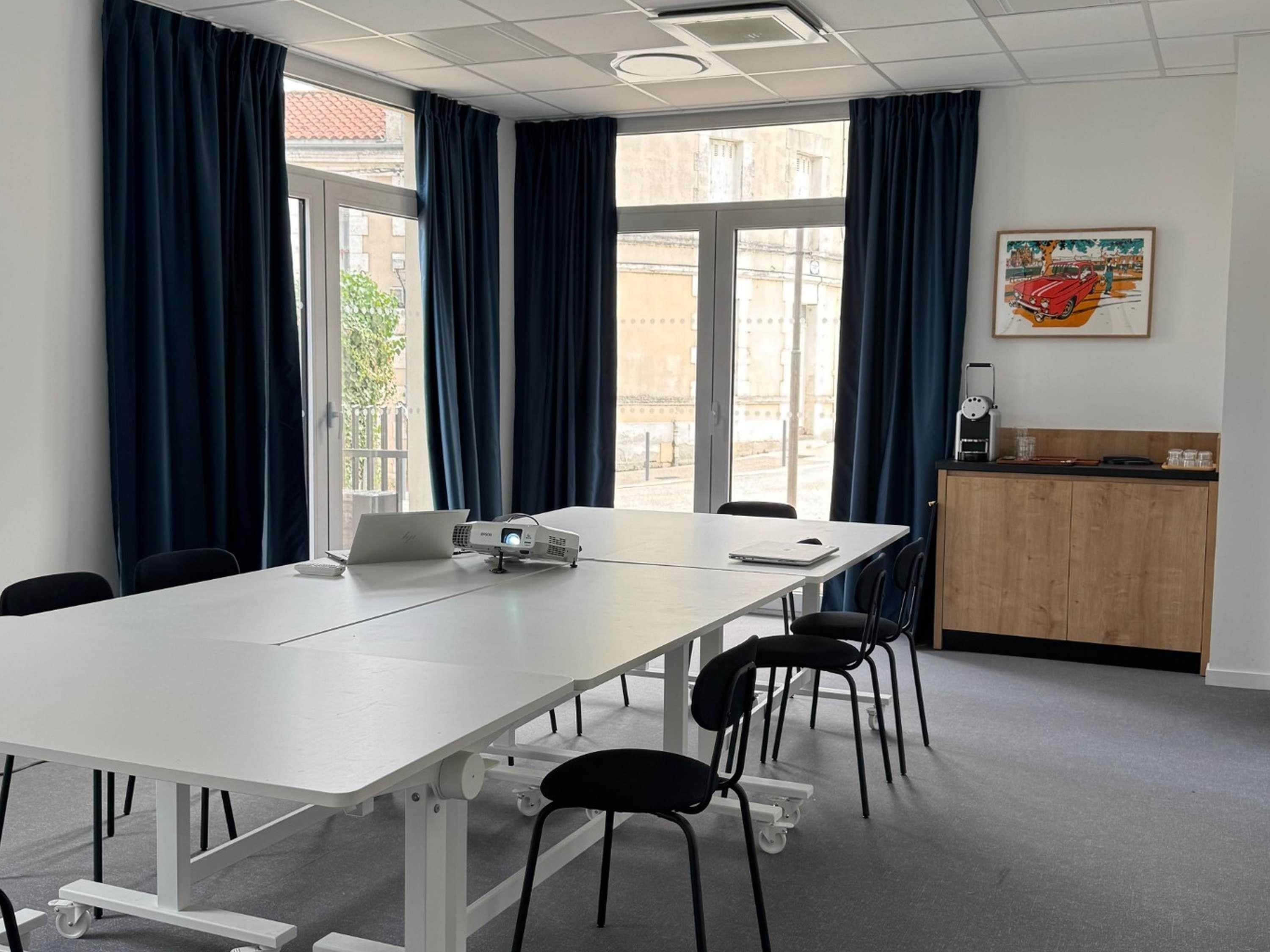 A conference room with a long table, chairs, a projector, a laptop, and a small kitchenette area.