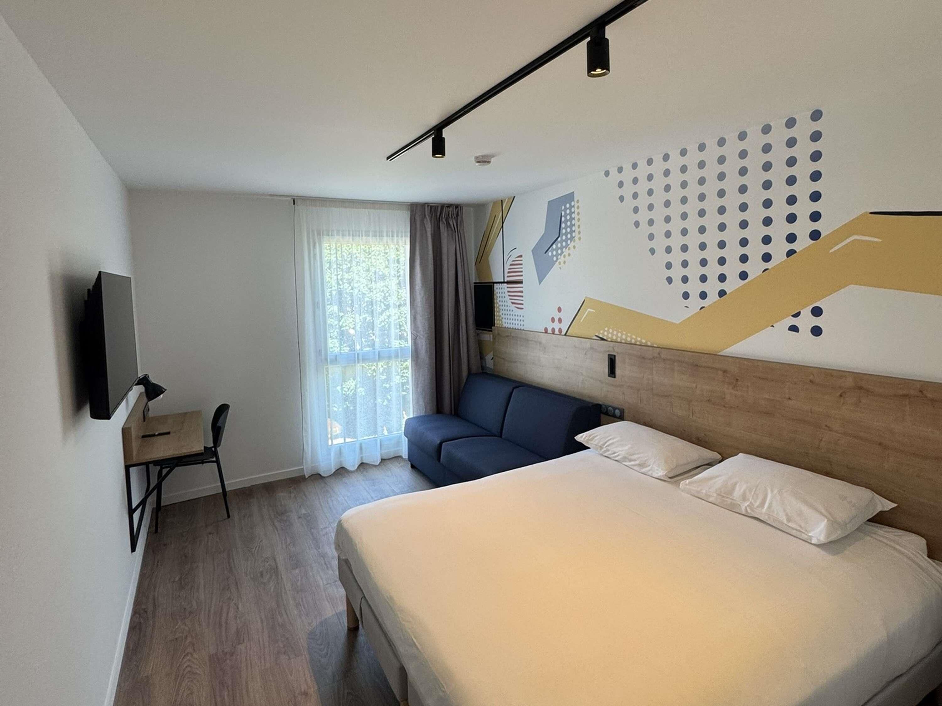 Habitación de hotel moderna con cama, escritorio, silla, televisor y ventana con cortinas.