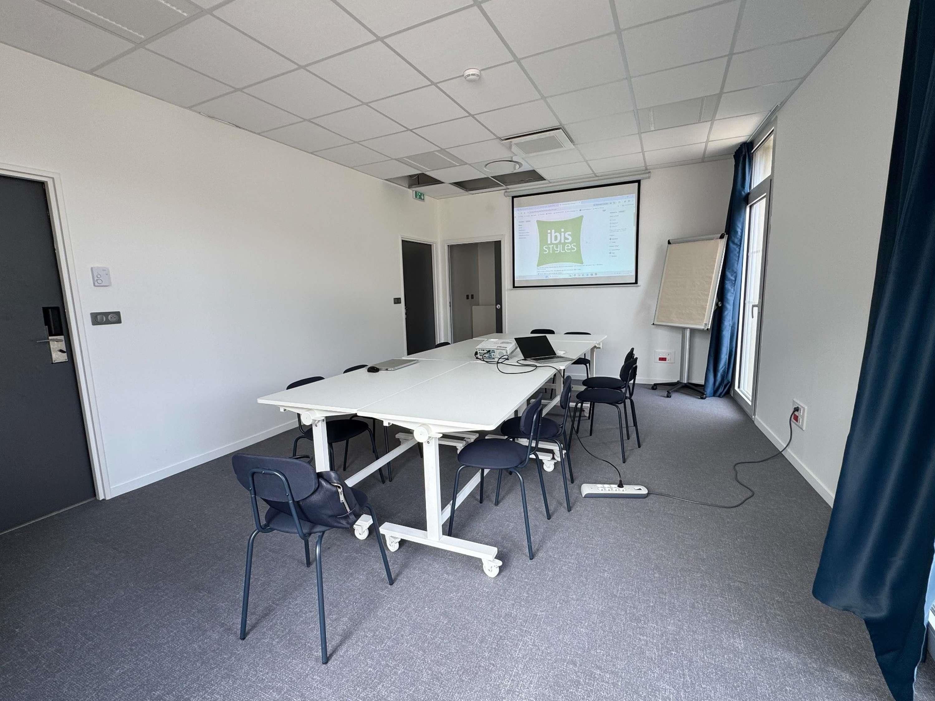 Uma sala de conferência com uma mesa branca, cadeiras, uma tela de projetor e um quadro branco.