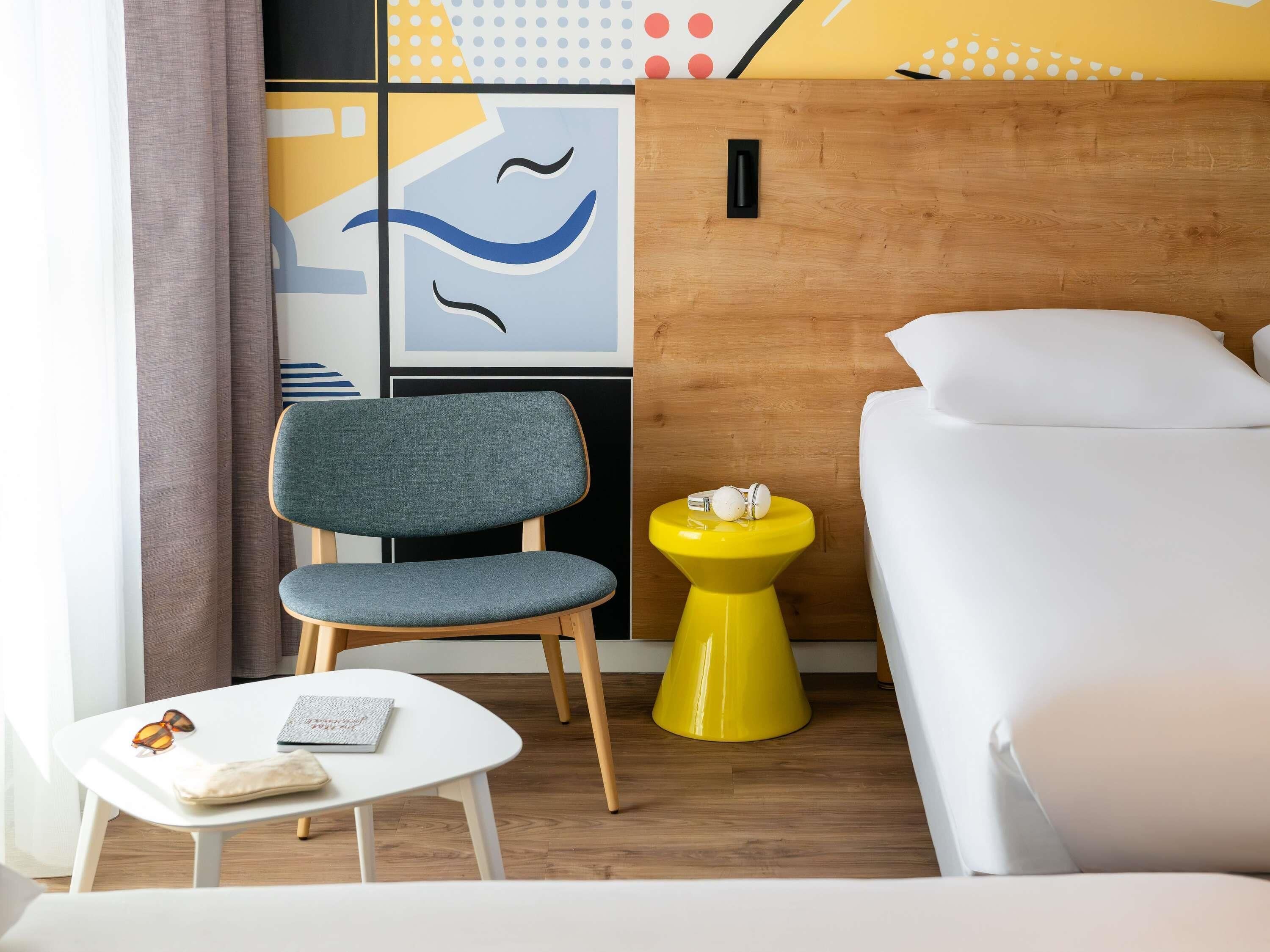 Une chambre d’hôtel avec un lit, une chaise, une petite table sur laquelle se trouvent un livre et des lunettes de soleil, et une table d’appoint jaune avec une boîte de mouchoirs.