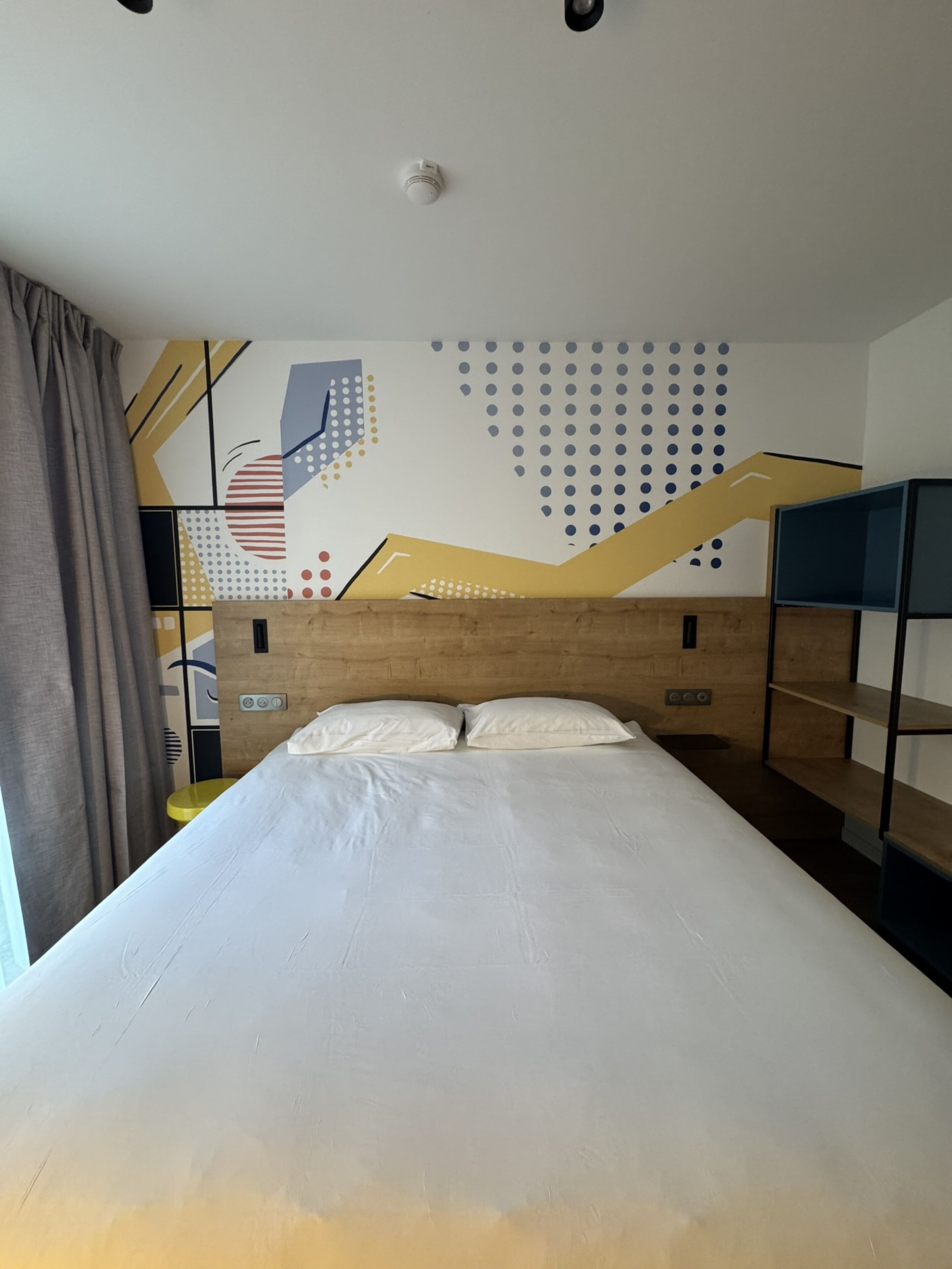 Habitación de hotel con cabecera de madera, cama con ropa blanca, pared decorada con arte abstracto y ventana con cortinas.