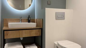 A modern bathroom with a round mirror, a white sink, a toilet, and a shelf with folded towels.