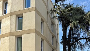 Um edifício de vários andares com fachada moderna, entrada visível e uma grande árvore na frente.