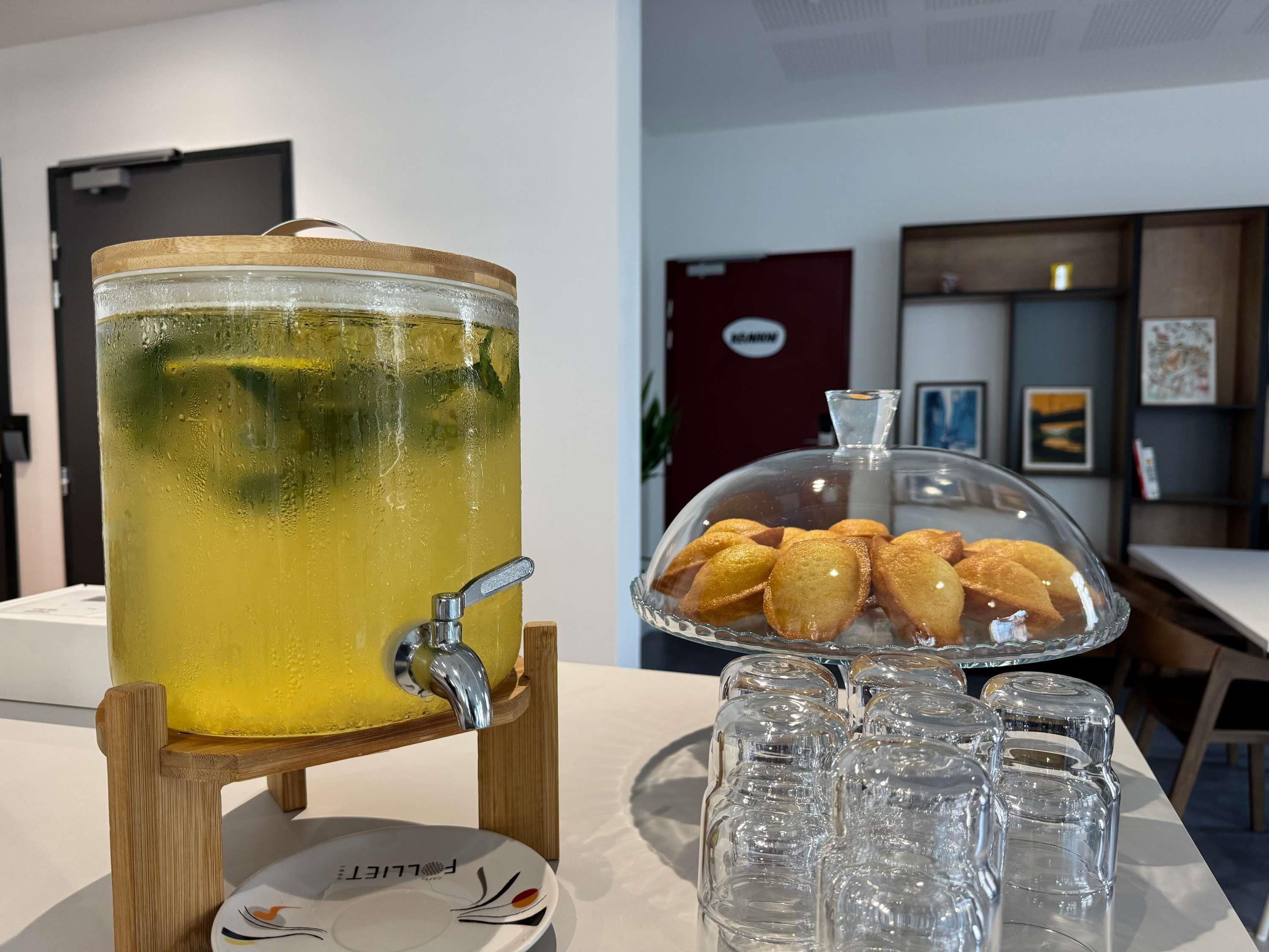 A beverage dispenser with a tap, a tray of pastries, and several glasses on a table.