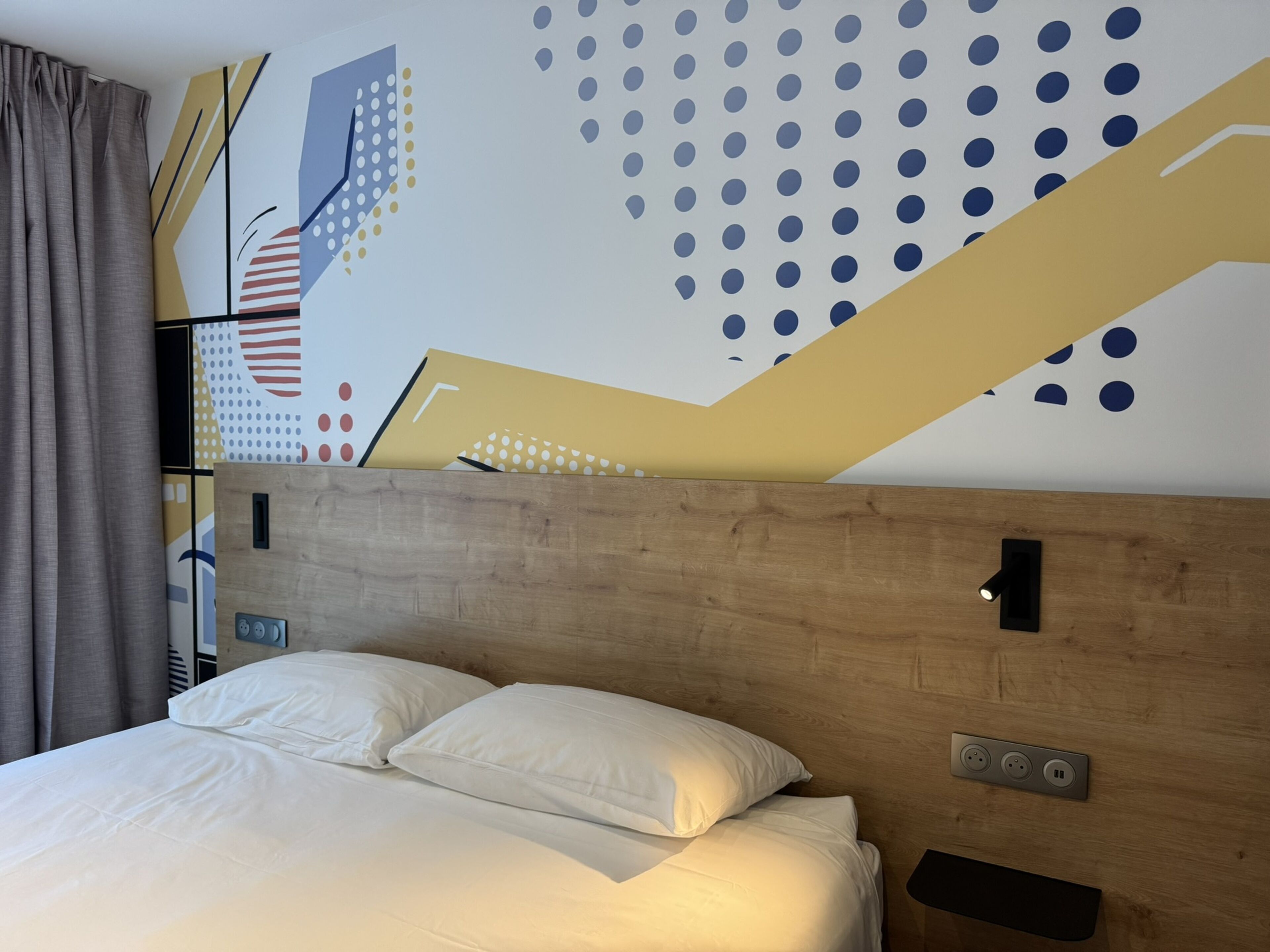 A modern hotel room with a wooden headboard, a bed with white linens, a wall-mounted light, and a power outlet.