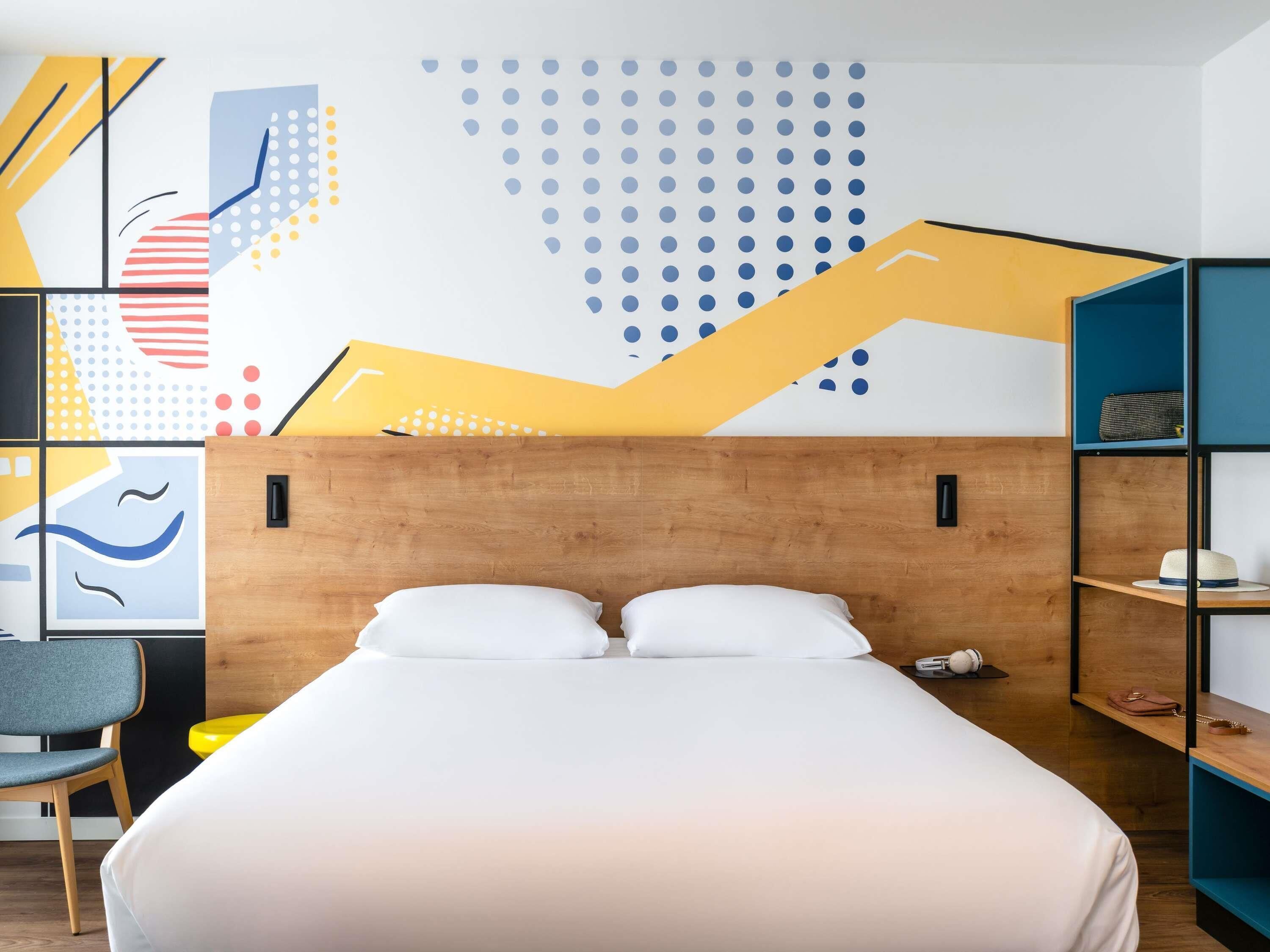 Habitación de hotel moderna con cabecera de madera, cama con sábanas blancas, escritorio con silla y una pared decorada con arte abstracto.