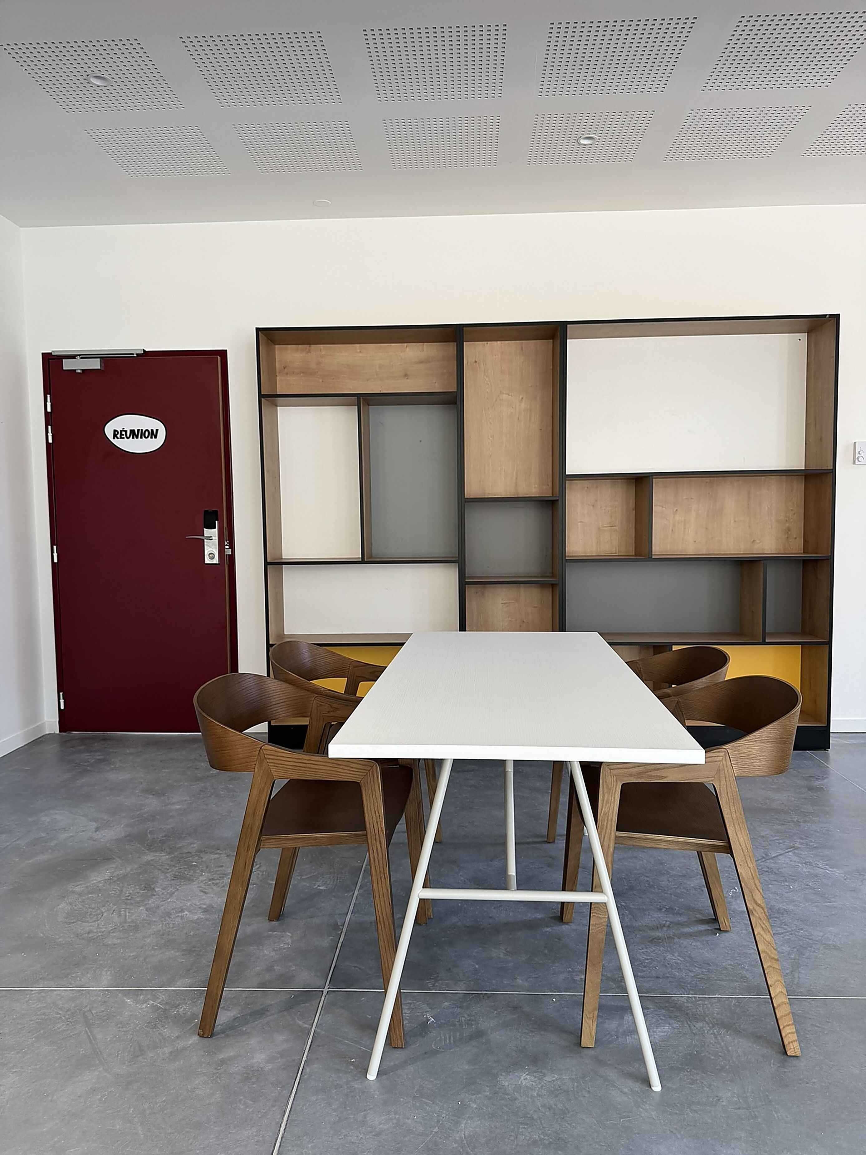 A conference room with a white table, wooden chairs, a red door labeled 'REUNION', and a built-in shelving unit.
