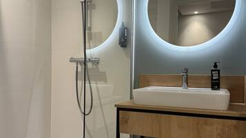 A modern bathroom with a glass-enclosed shower, a wooden vanity with a sink, and a mirror with a circular light.