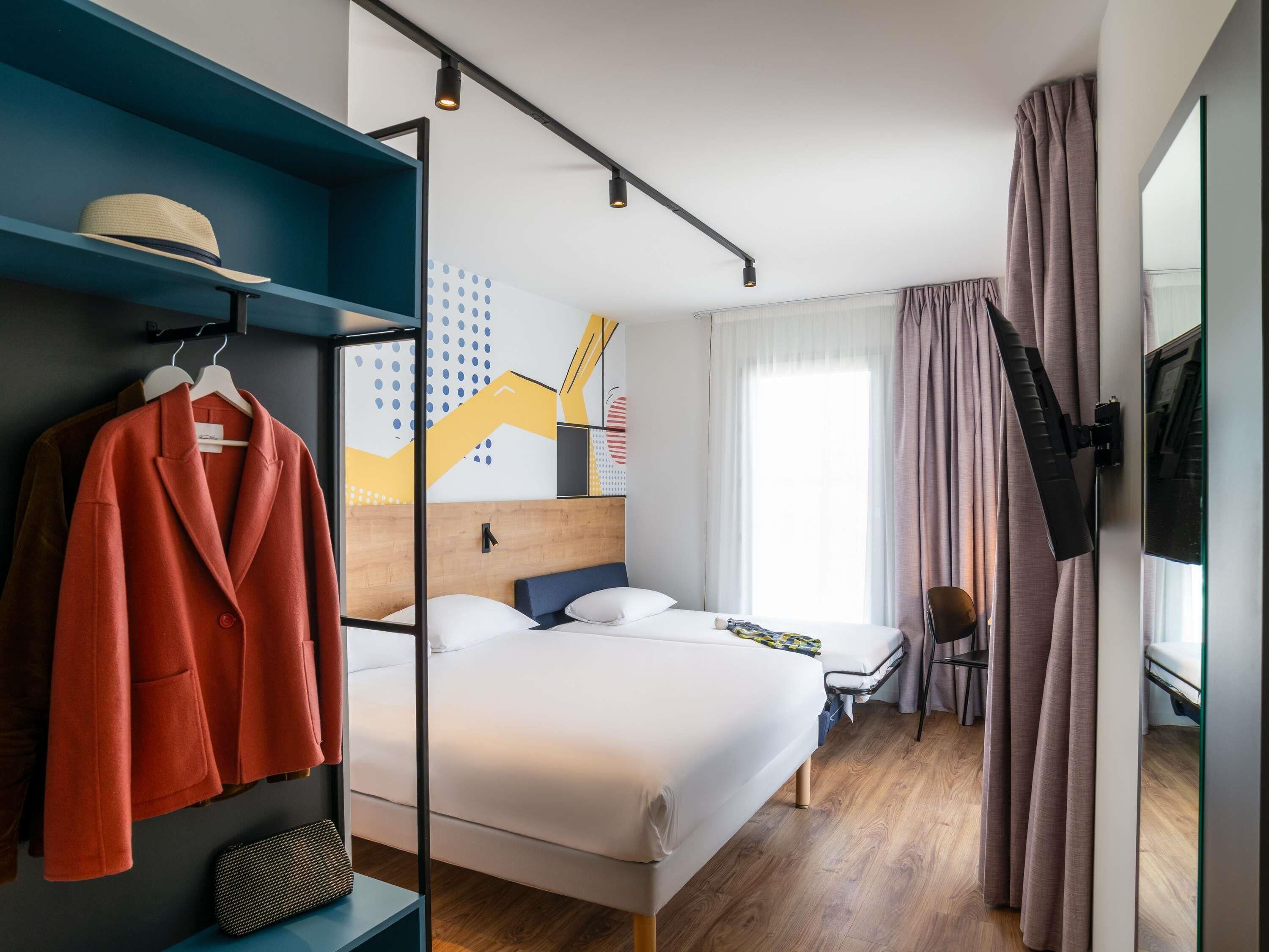 Quarto de hotel com cama, escrivaninha, cadeira, televisão e guarda-roupa.