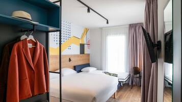 Quarto de hotel com cama, escrivaninha, cadeira, televisão e guarda-roupa.