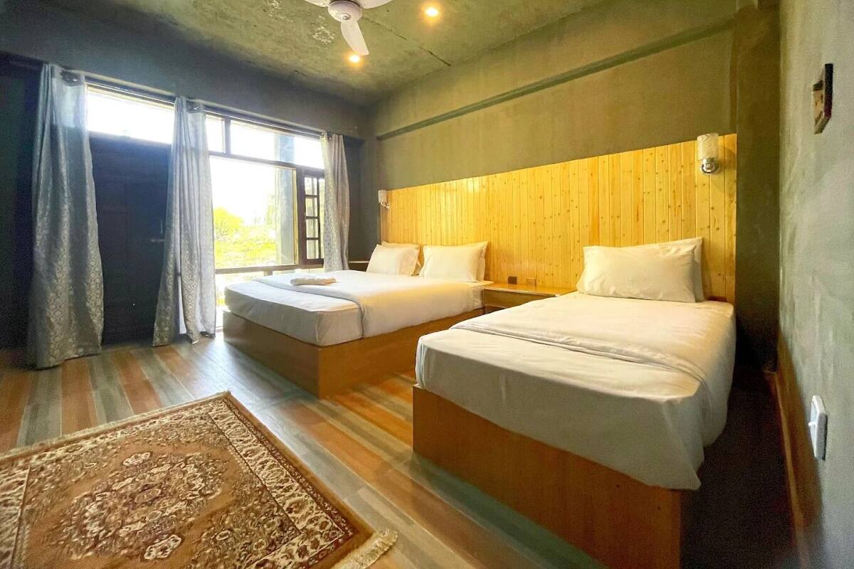 Habitación de hotel con dos camas, ventilador de techo y un amplio ventanal con cortinas.