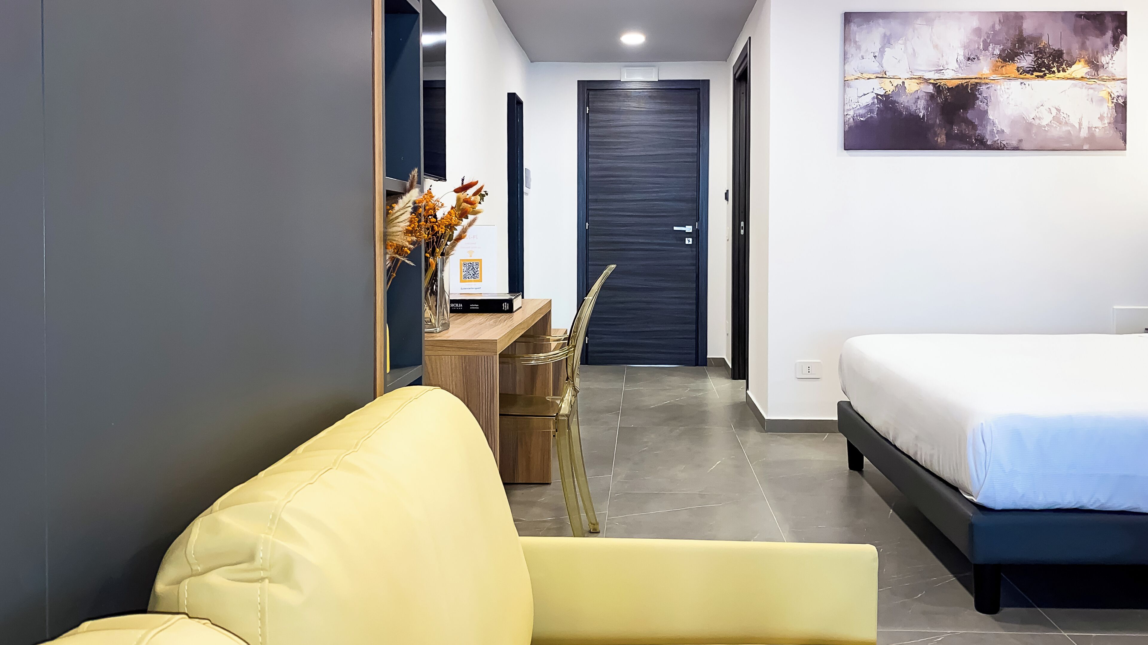 A modern hotel room with a bed, a sofa, a desk with a vase of flowers, and a door.