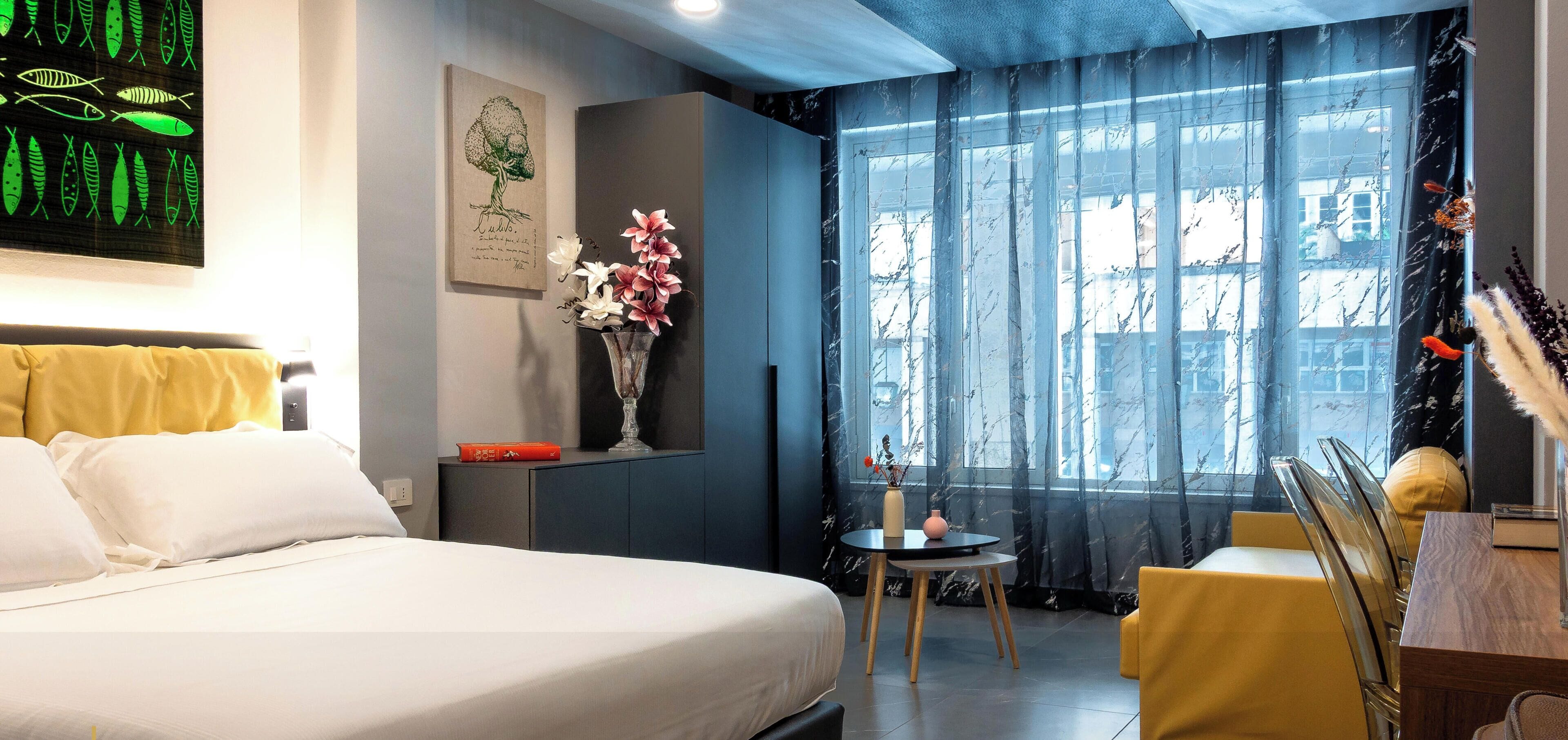 A hotel room with a bed, a small table with chairs, a vase with flowers, a painting, and a window with curtains.