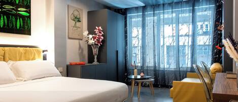 A hotel room with a bed, a small table with chairs, a vase with flowers, a painting, and a window with curtains.