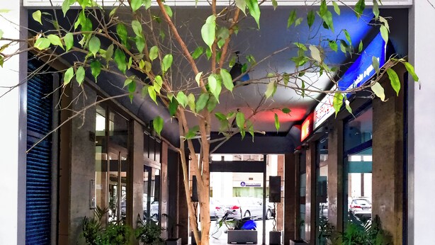 A covered walkway with potted plants, a blue sign, and a visible car in the background. - Suiteincenter Rooms (Palermo)