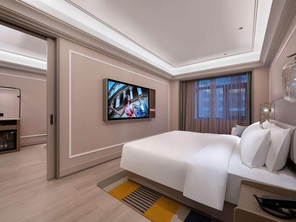 A modern hotel room with a large bed, a flat-screen TV mounted on the wall, and a bedside table with a lamp.