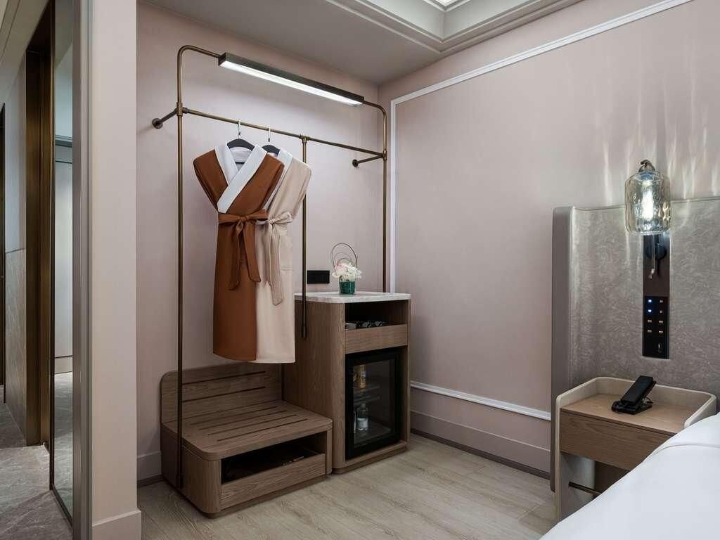 A hotel room with a bed, a bedside table with a phone, a small wooden bench, a coat rack with clothes, and a wall-mounted light fixture.