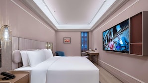 A hotel room with a bed, bedside table, lamp, TV, and a small desk with chair.