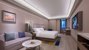 A hotel room with a bed, a sofa, a small round table with fruit, a desk, and a TV.