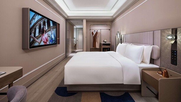 A modern hotel room with a large bed, a TV mounted on the wall, a desk, and a chair.