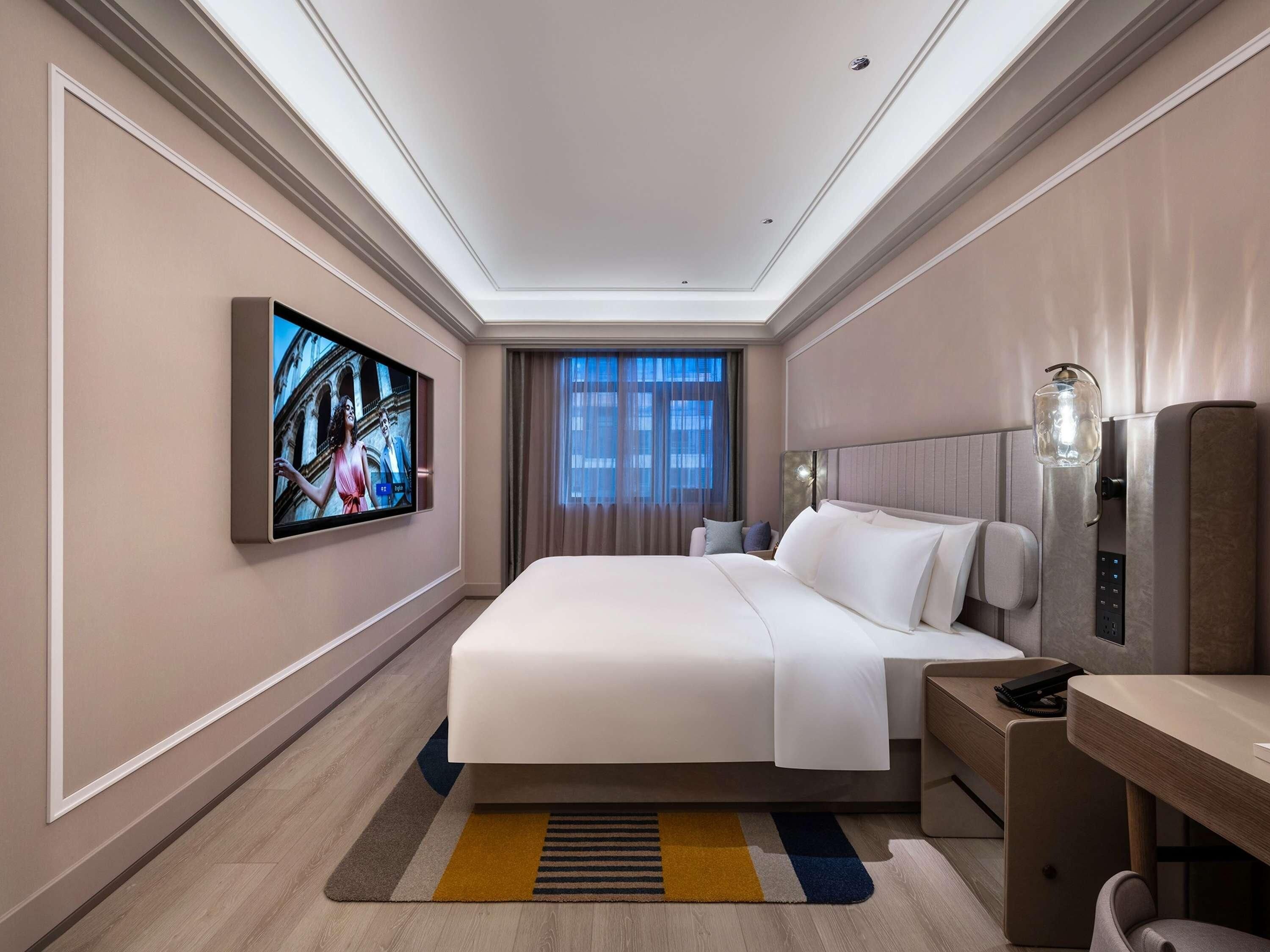 A hotel room with a bed, a desk, a television mounted on the wall, and a window with curtains.