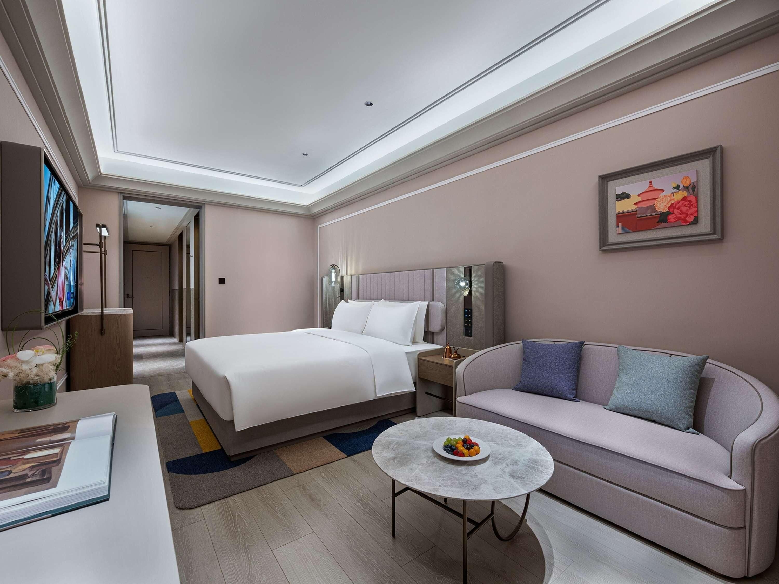Une chambre d’hôtel avec un lit, un canapé, une table ronde avec des fruits, une télévision et un tableau au mur.