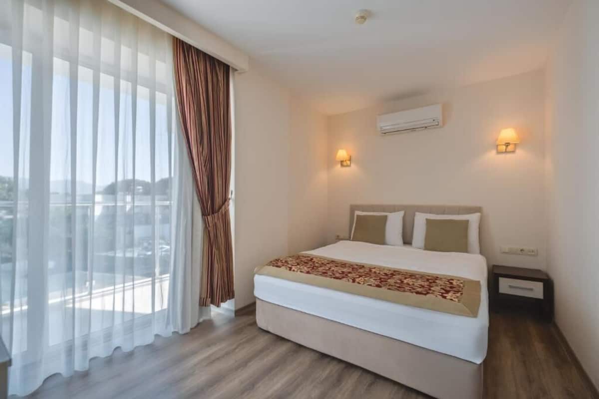 A hotel room with a bed, curtains, two wall-mounted lights, an air conditioning unit, and a view through the window.